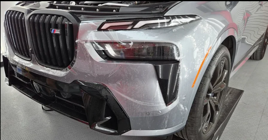 Close-up of a silver BMW SUV with partial paint protection film being applied to the front bumper and fender area, showing air bubbles and edges still being worked on.