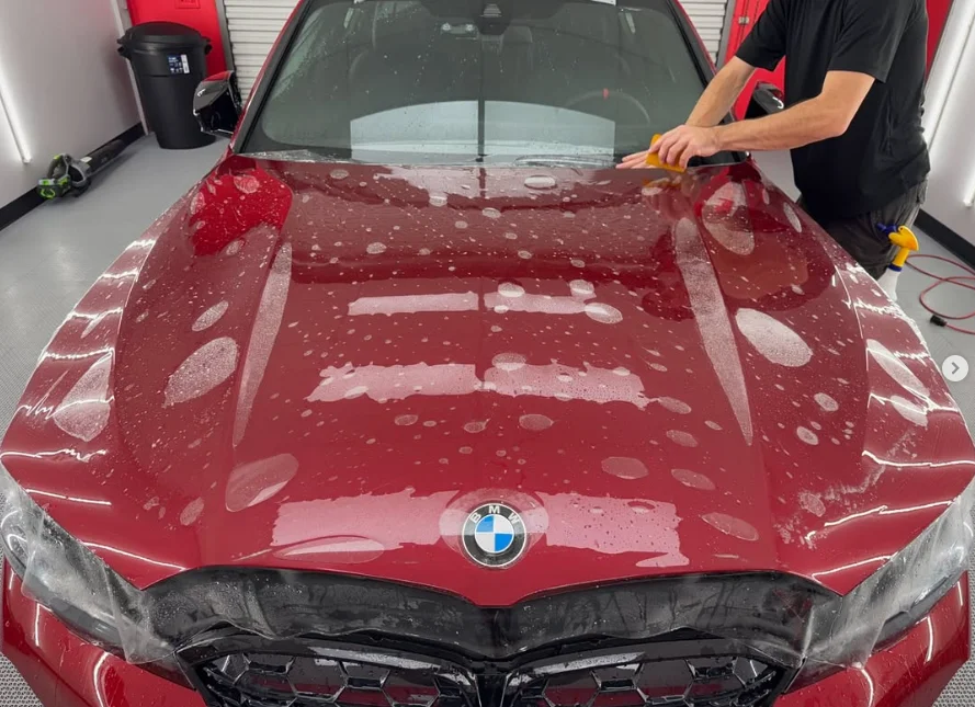 Red BMW sedan with paint protection film being applied to the hood, showing bubbles and moisture as a detailer smooths the surface with a squeegee.