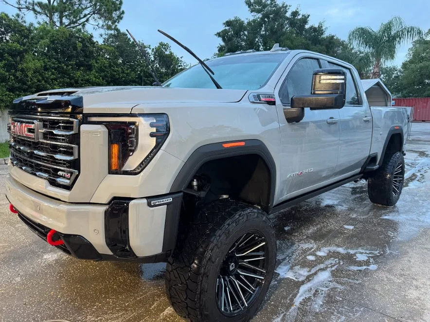 Lifted white GMC Sierra 2500HD AT4 with custom black wheels and red tow hooks, parked outdoors on a wet surface after a wash.