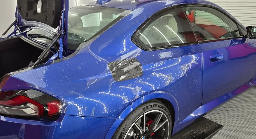 Blue BMW coupe with paint protection film being applied to the rear fender, showing wet application and partially installed material.