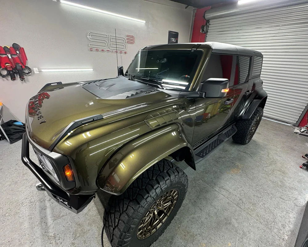Olive green Ford Bronco with matte black accents and off-road tires, parked inside a detailing studio under LED lights.