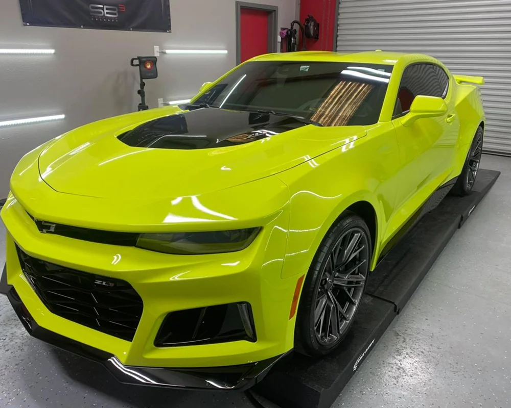 Bright yellow-green Chevrolet Camaro ZL1 with a black hood scoop, parked indoors on a detailing platform under LED lighting.