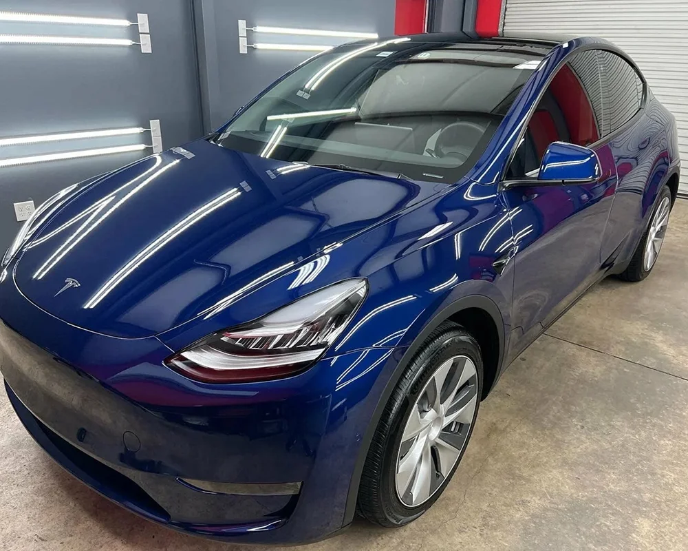 Shiny deep blue Tesla Model Y parked in a detailing studio under bright LED lights, showcasing a flawless exterior finish and clean glass surfaces.