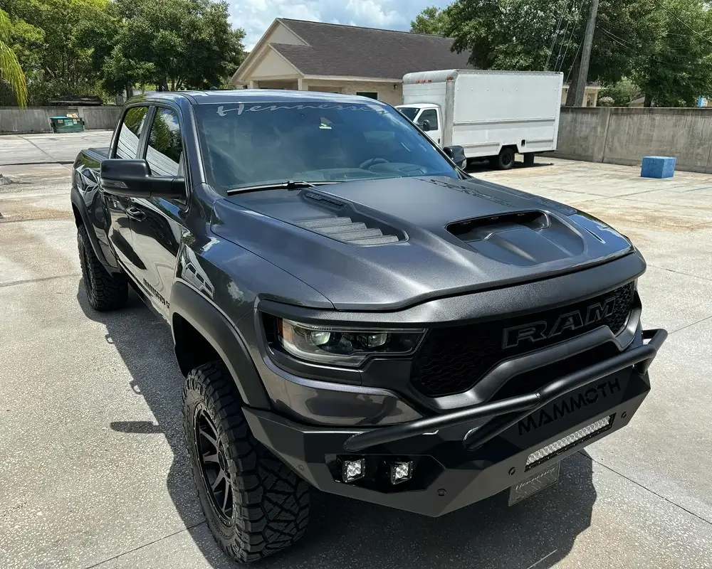 Matte black RAM 1500 TRX Hennessey Mammoth edition truck with aggressive off-road tires and custom bumper, parked in an outdoor lot under bright sunlight.