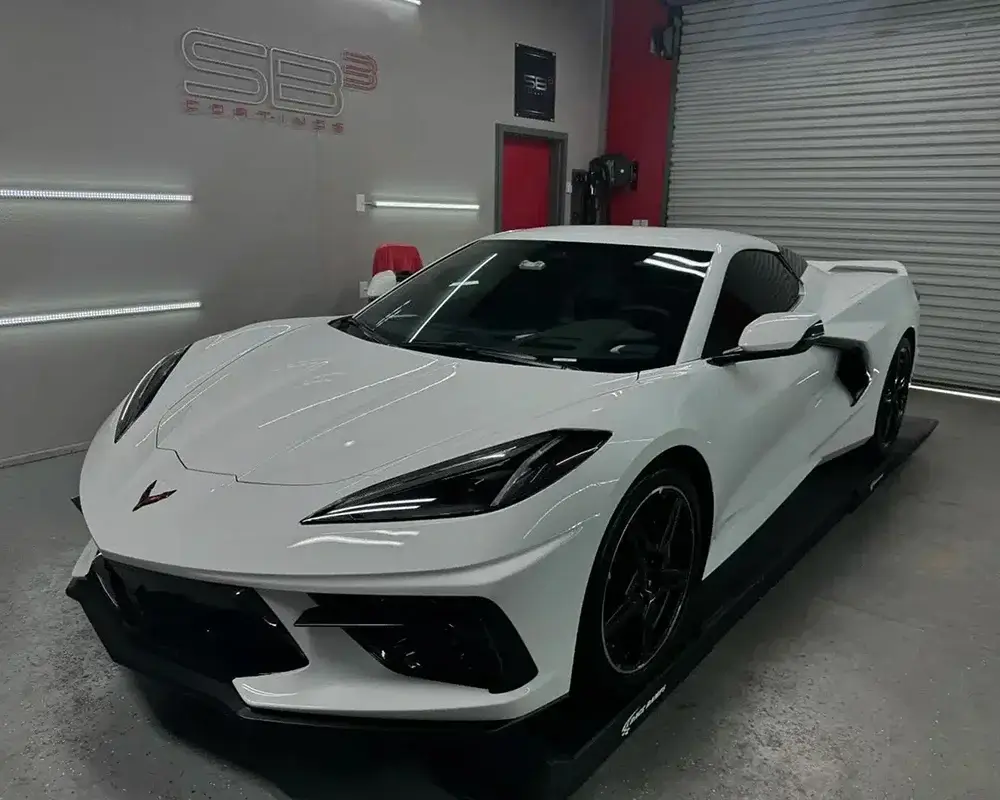 White Chevrolet Corvette C8 with black wheels and dark-tinted windows, positioned on a detailing platform under studio lighting.